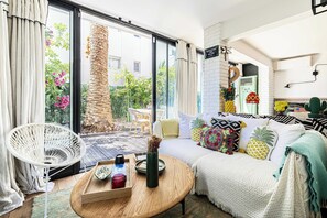 A modern living room with a sliding glass door to a garden, a white chair, a sofa with colorful pillows, and a wooden coffee table with decorative items. - Charming Apartment With Terrace a/c 4P - Cannes (Cannes)
