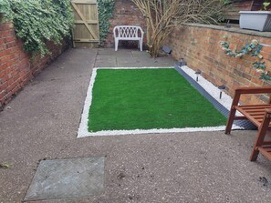 A small backyard with a green artificial turf, a white plastic bench, a brick wall, and a wooden gate. - Bentley View,2 bed House Crewe (Crewe)