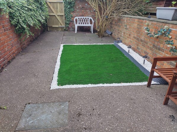A small backyard with a green artificial turf, a white plastic bench, a brick wall, and a wooden gate. - Bentley View,2 bed House Crewe (Crewe)