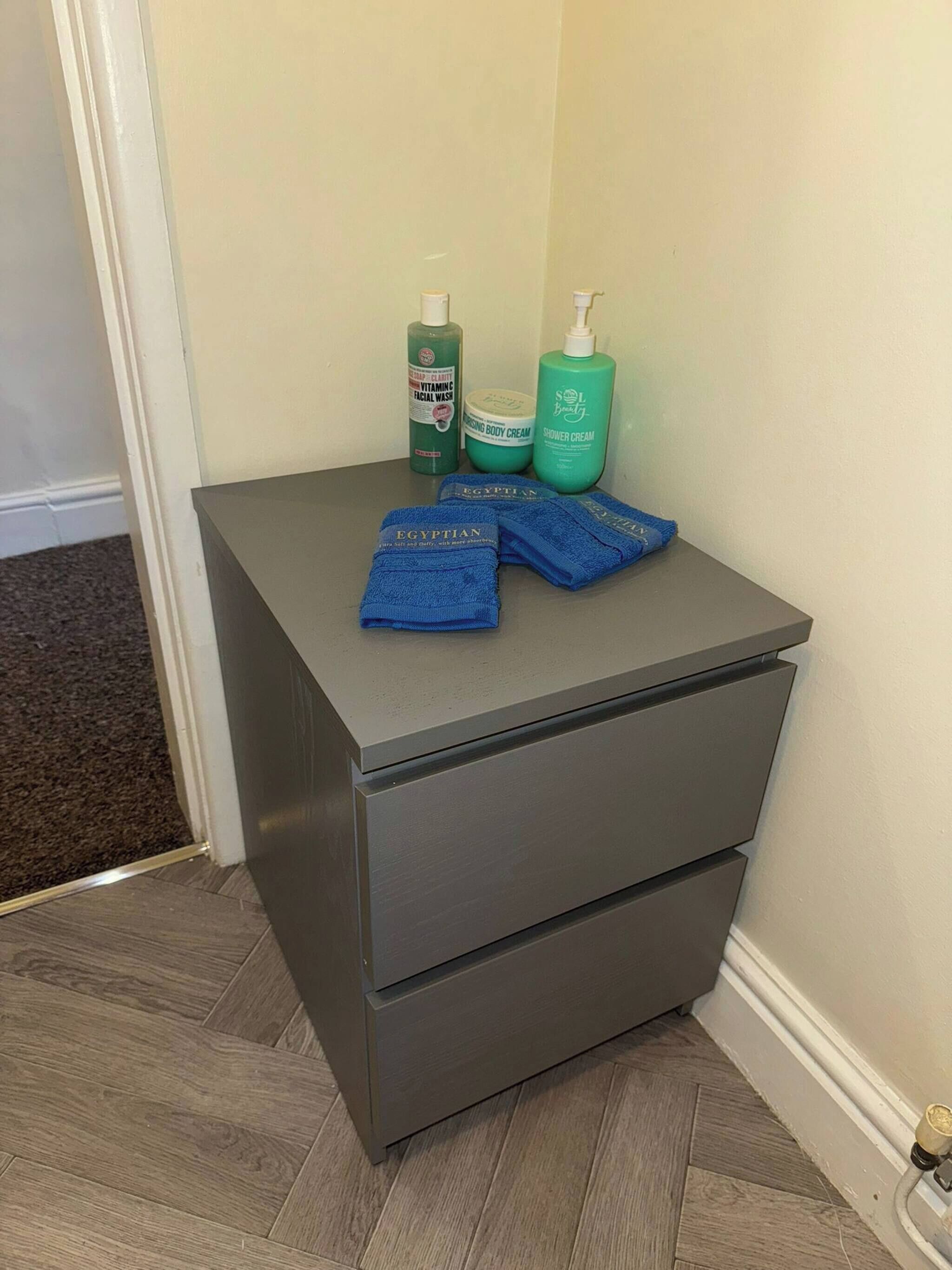A small gray bedside table with two drawers, a bottle of green facial wash, a bottle of body cream, a bottle of shower cream, and two blue towels.