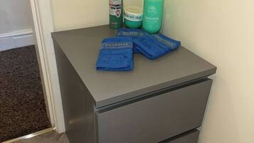 A small gray bedside table with two drawers, a bottle of green facial wash, a bottle of body cream, a bottle of shower cream, and two blue towels.