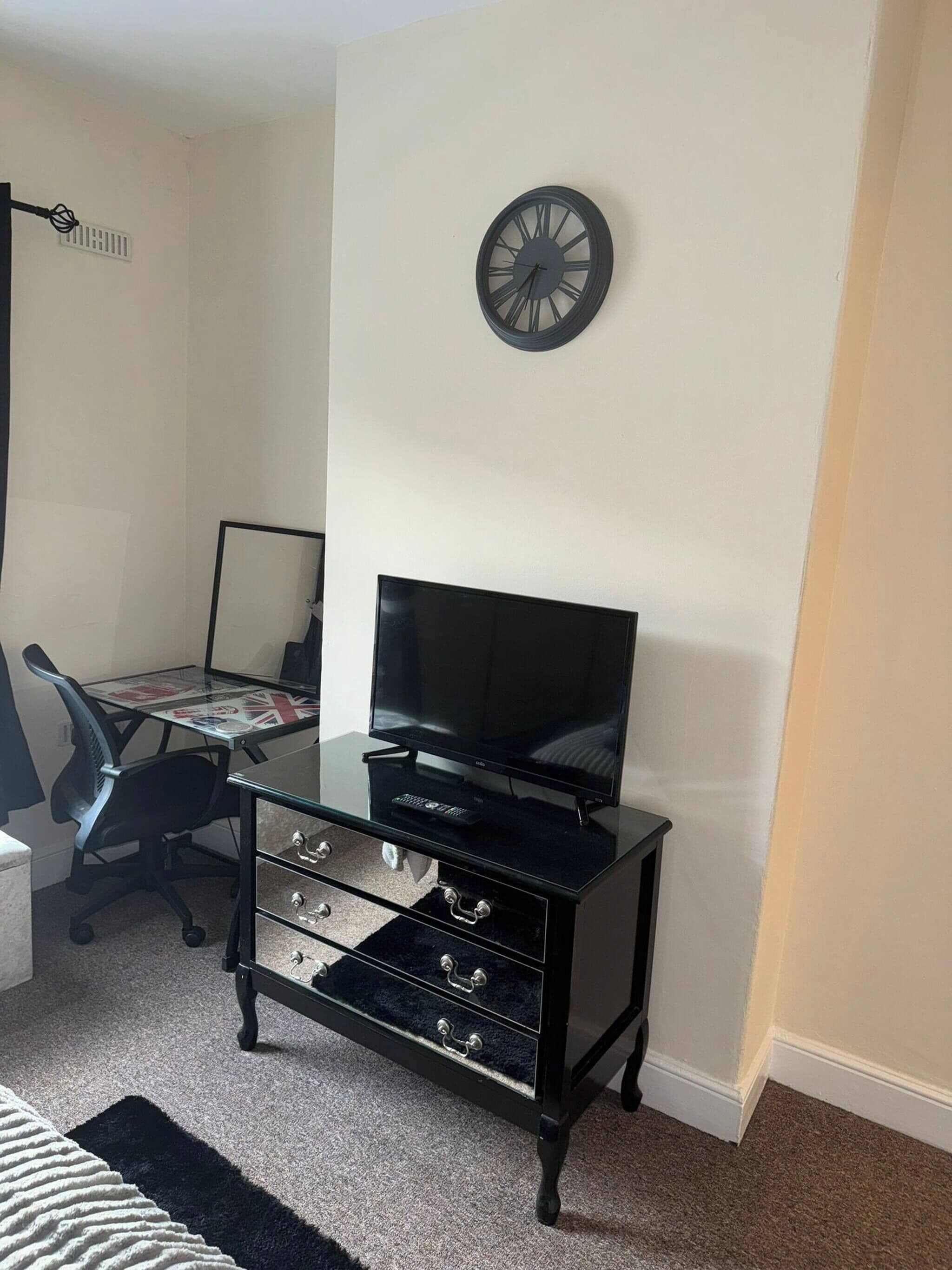 Quarto com um rack preto para TV, uma televisão de tela plana, uma mesa com cadeira e um relógio de parede.