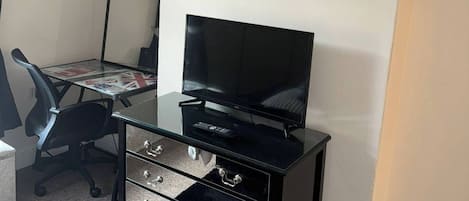 Une pièce comprenant un meuble TV noir, un téléviseur à écran plat, un bureau avec une chaise et une horloge murale.