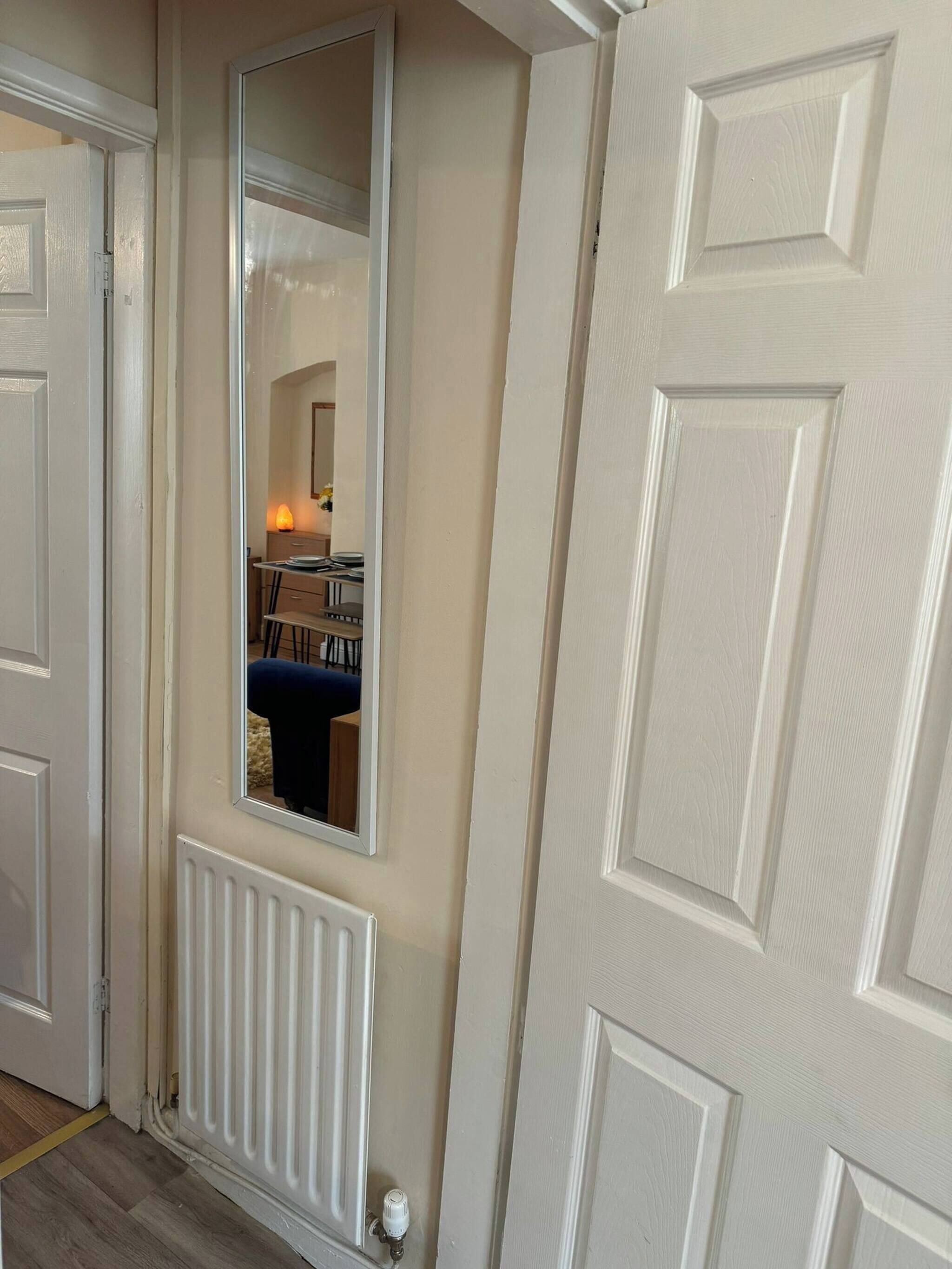 A room with a white door, a radiator, and a mirror reflecting a blue chair and a table with a lamp.