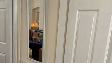 A room with a white door, a radiator, and a mirror reflecting a blue chair and a table with a lamp.