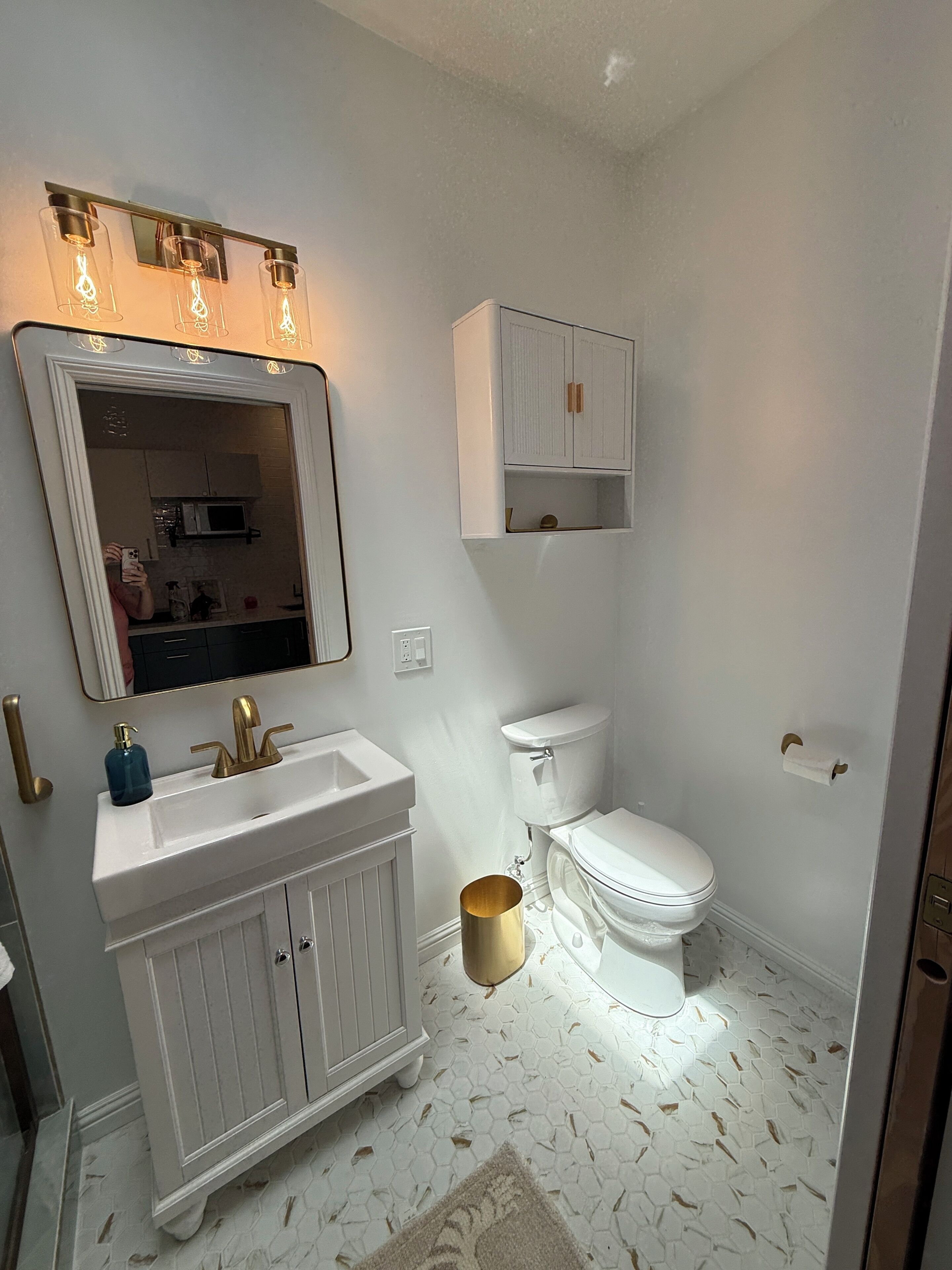A bathroom with a white vanity, a toilet, a cabinet above the toilet, and a mirror with lights.
