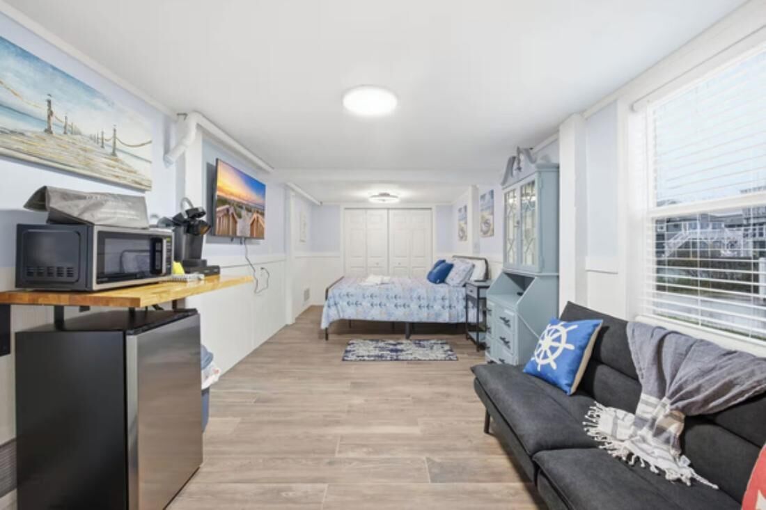 A compact room with a kitchenette, a bed, a sofa, and a window with blinds.
