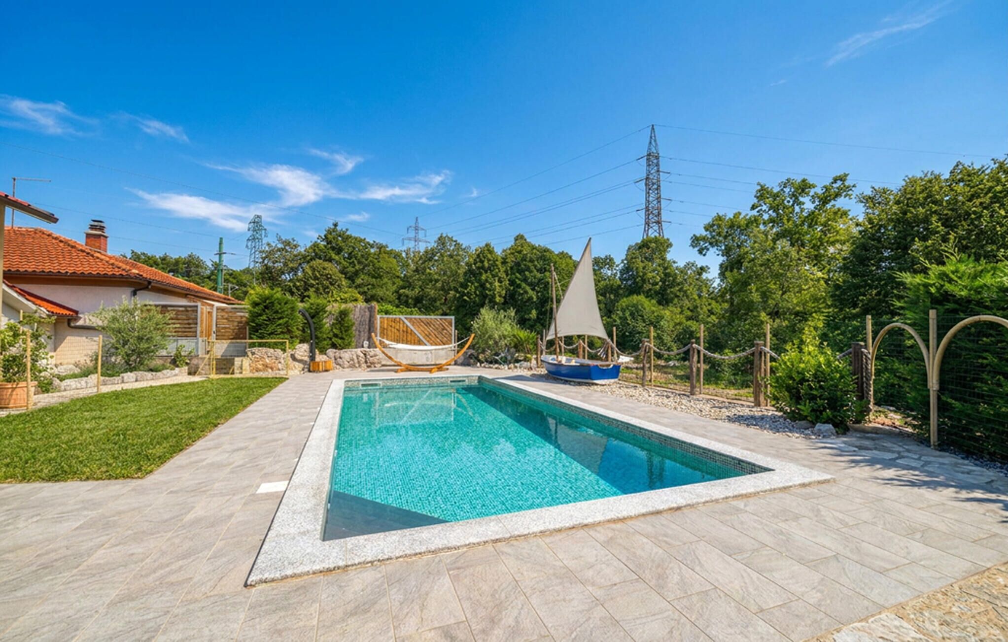 En swimmingpool med ett kaklat poolområde, omgiven av grönska och ett hus med ett rött tak.