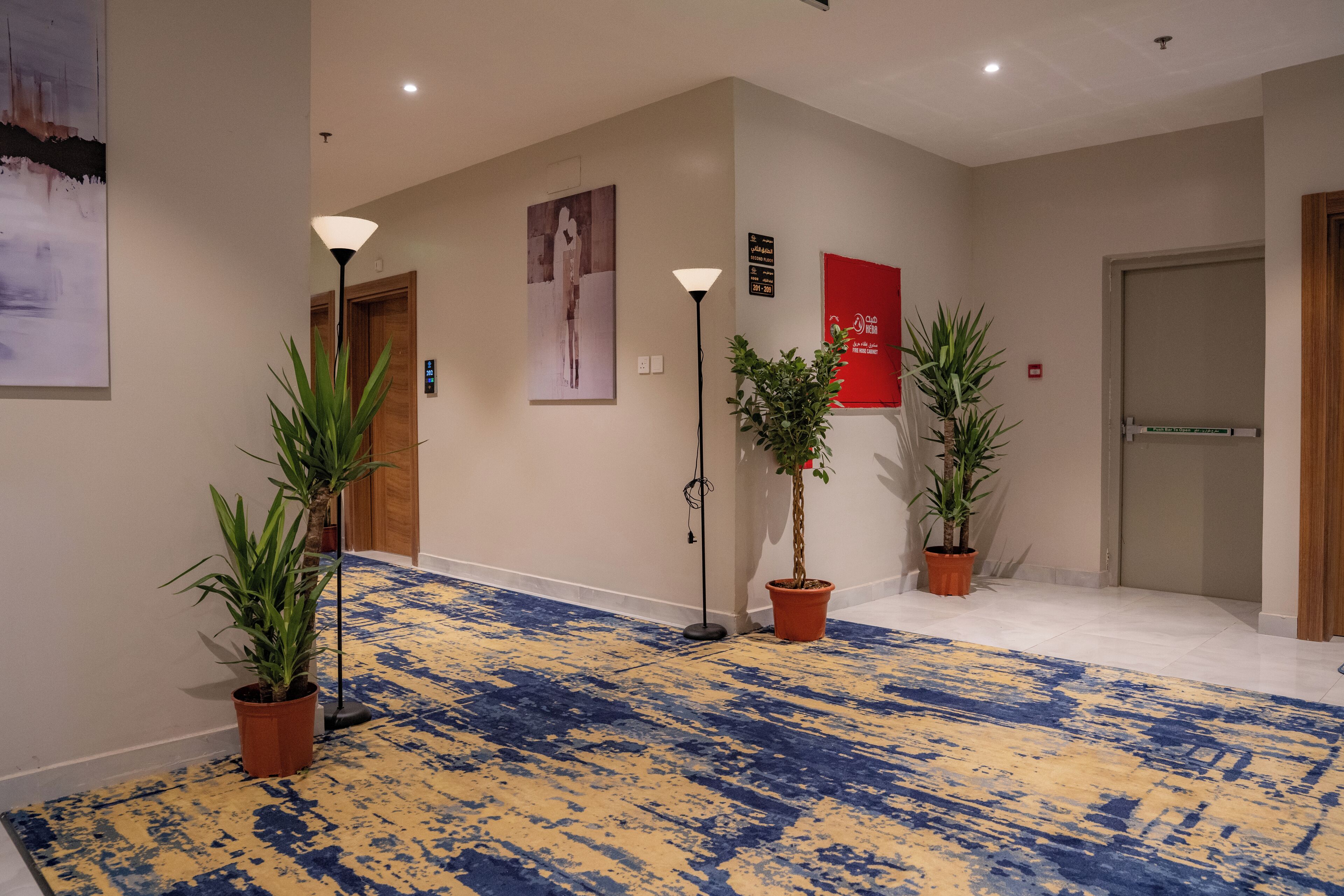 A hotel hallway with a blue and yellow patterned carpet, potted plants, a wall-mounted artwork, and a sign indicating the second floor.