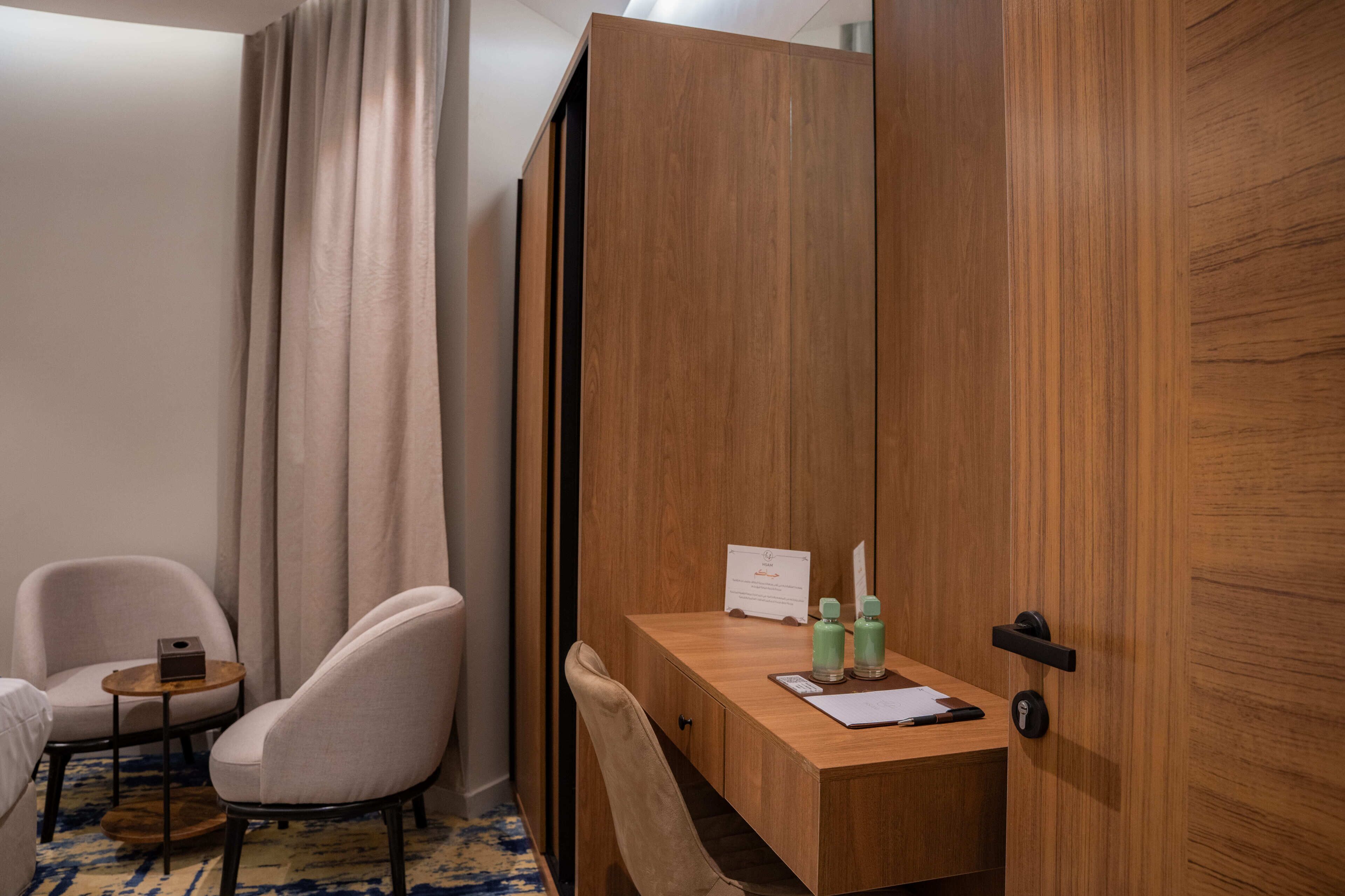 Hotellihuone, jossa on puinen työpöytä, kaksi tuolia, pieni pöytä, verho ja ovi.