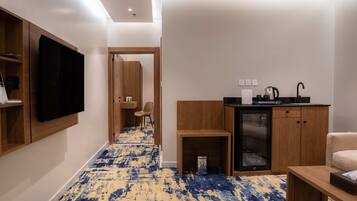 A hotel room with a TV, a kitchenette, a small table, a chair, and a carpeted floor.