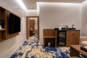 A hotel room with a TV, a kitchenette, a small table, a chair, and a carpeted floor.