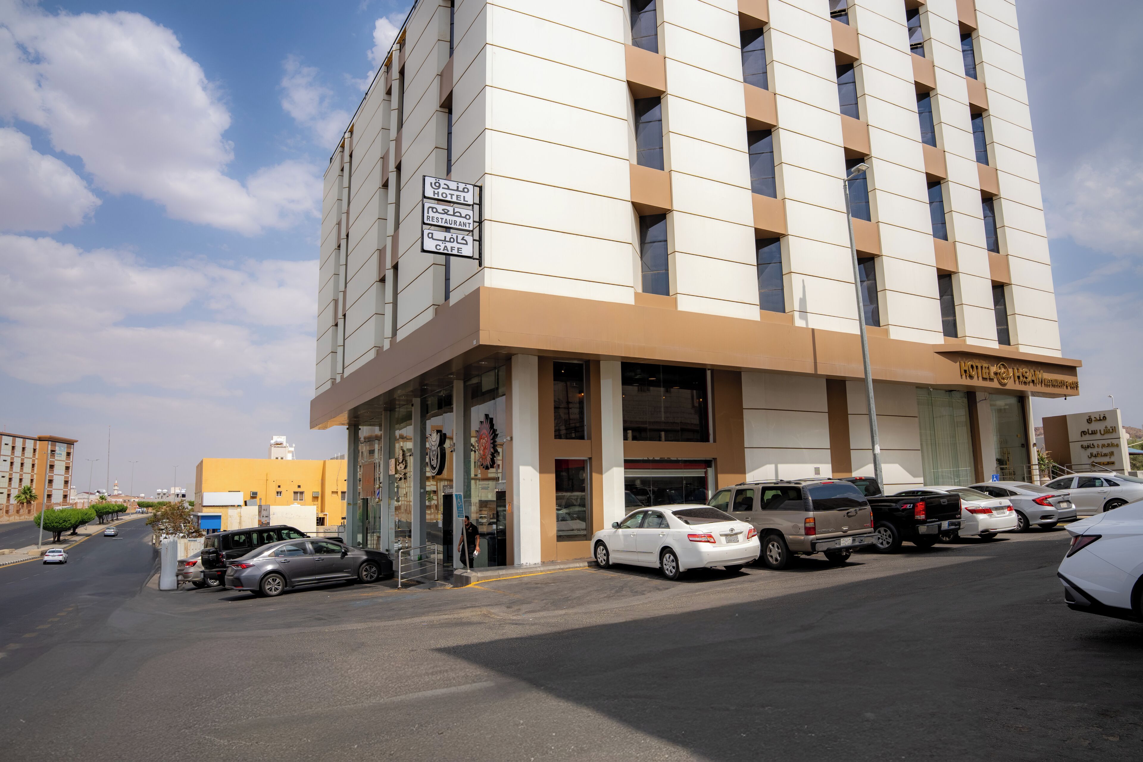 Un hôtel à plusieurs étages doté d’une façade moderne, un parking où sont garées plusieurs voitures et un ciel dégagé.