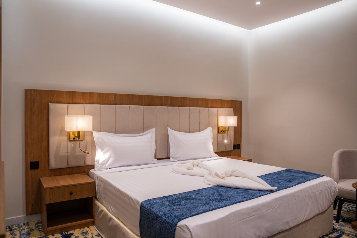 Habitación de hotel con una cama grande, dos lámparas de noche, un cabecero de madera, una mesita de noche y una silla.