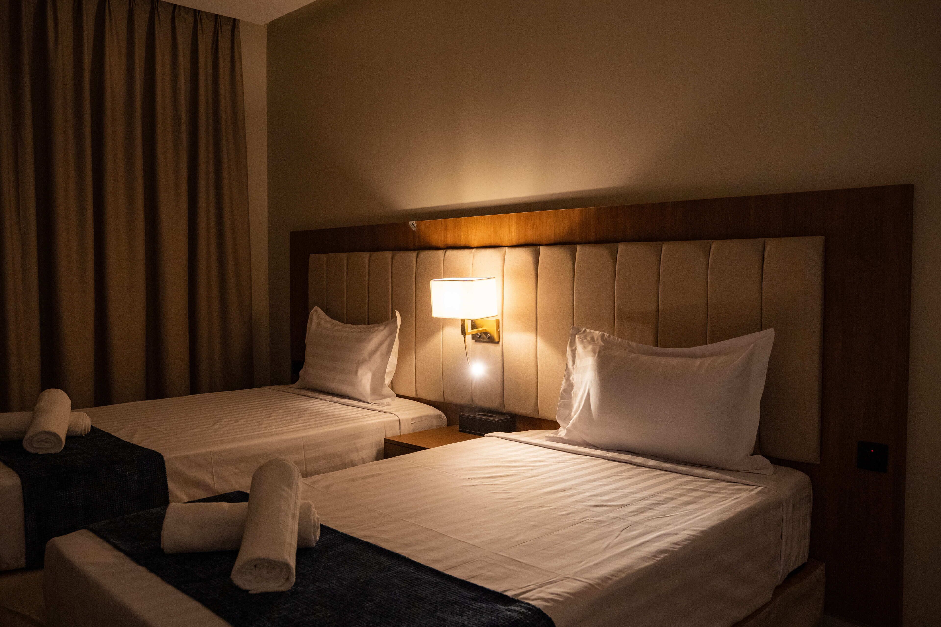 A hotel room with two beds, a headboard, a nightstand, a lamp, and a curtain.