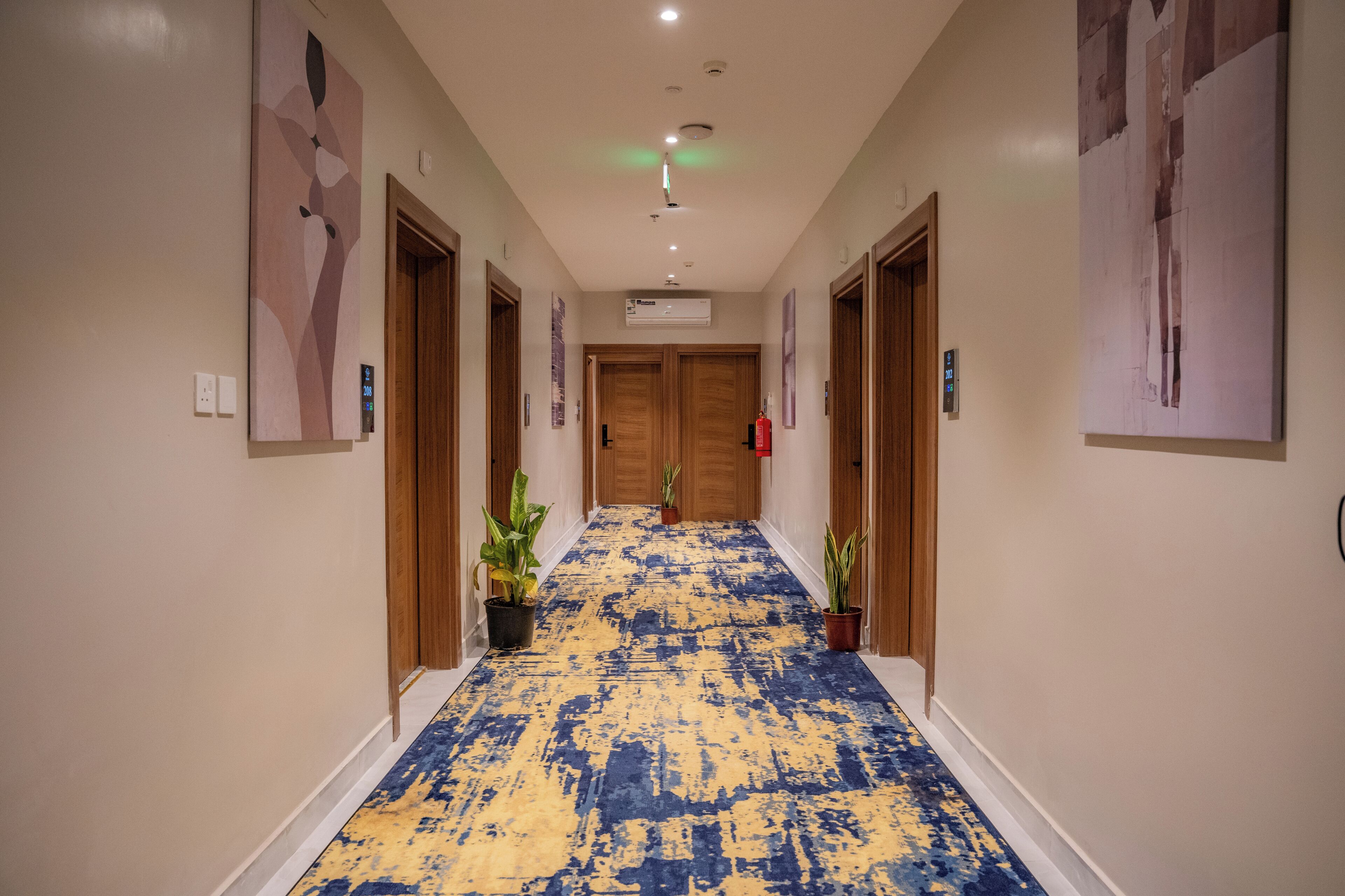 Corredor de hotel com carpete estampado em azul e amarelo, portas de madeira e plantas em vasos.