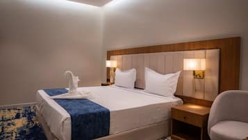 Habitación de hotel con una cama grande, dos lámparas de noche, cabecera de madera, alfombra con motivos azules y amarillos, y una sola silla.