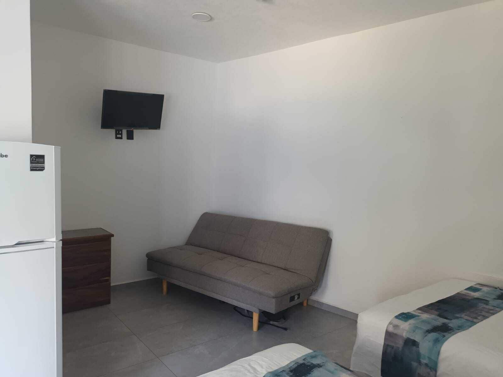 Una habitación pequeña y escasamente amoblada con un sofá, una cama, un televisor montado en la pared y un refrigerador blanco.