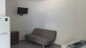 Una habitación pequeña y escasamente amoblada con un sofá, una cama, un televisor montado en la pared y un refrigerador blanco.