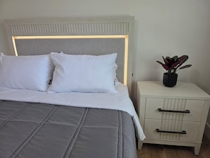 Un lit avec une tête de lit, deux oreillers, une table de chevet avec des tiroirs et une plante en pot.