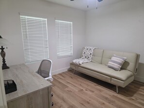 Una habitación con escritorio, dos ventanas con persianas, un sofá con cojines estampados y una silla.
