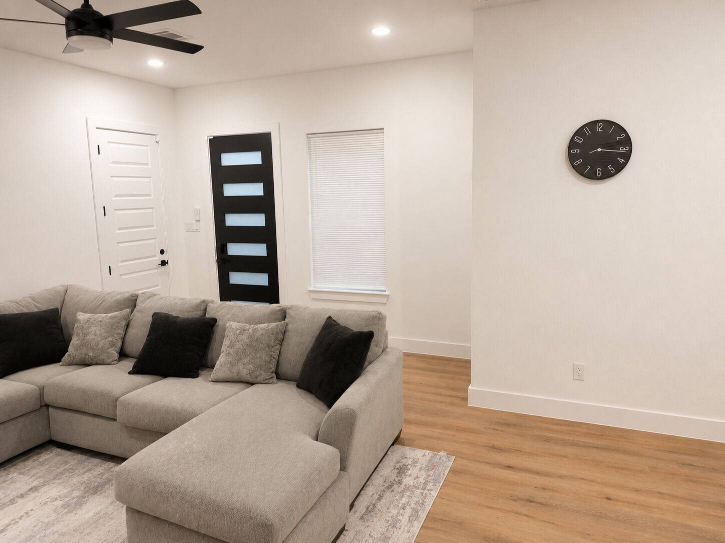 A living room with a sectional sofa, a ceiling fan, a clock on the wall, and a door with frosted glass panels.
