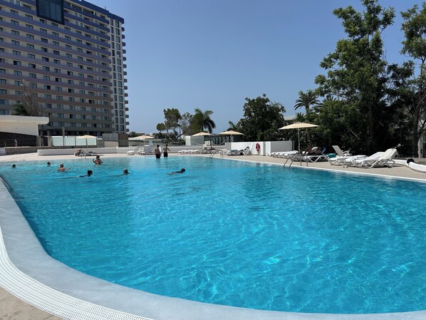 En swimmingpool med solstolar och parasoller, omgiven av en byggnad och träd.