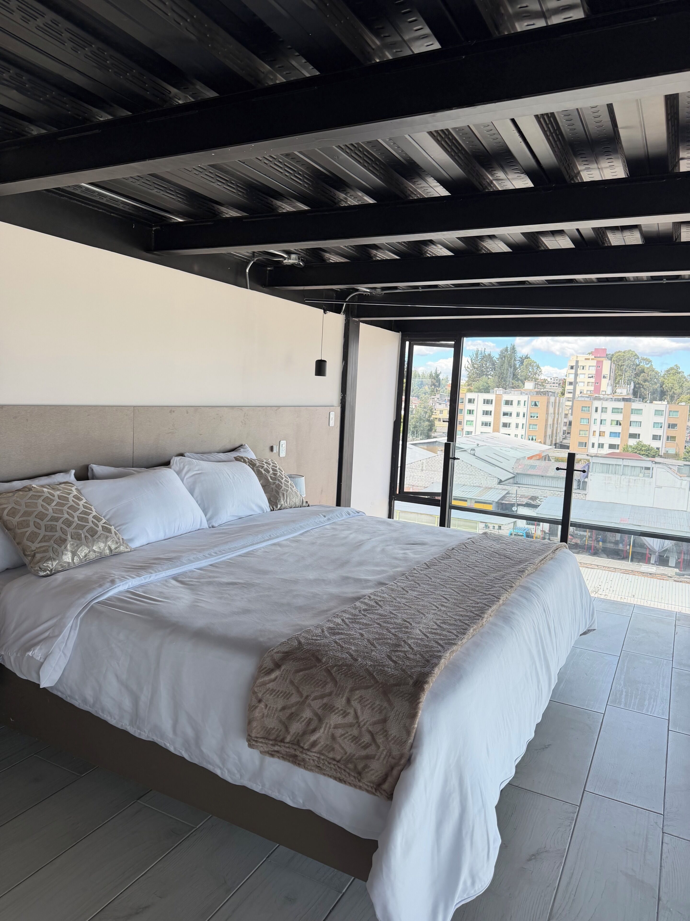 Ein modernes Schlafzimmer mit einem großen Bett, Blick auf die Stadt durch ein großes Fenster und einer Decke mit freiliegenden Balken.