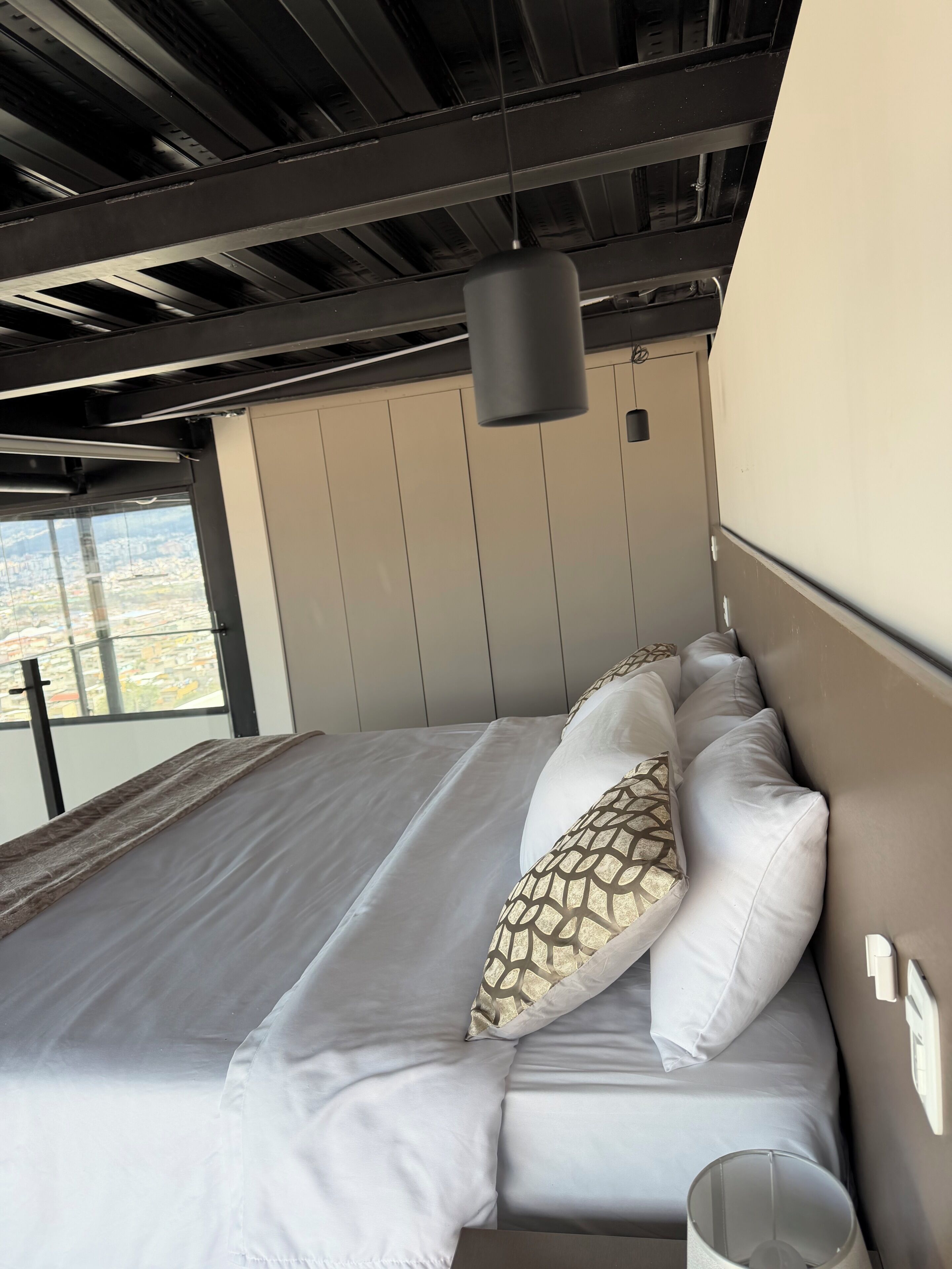 A bed with white linens and patterned pillows, a hanging light fixture, and a view of the city through a window.