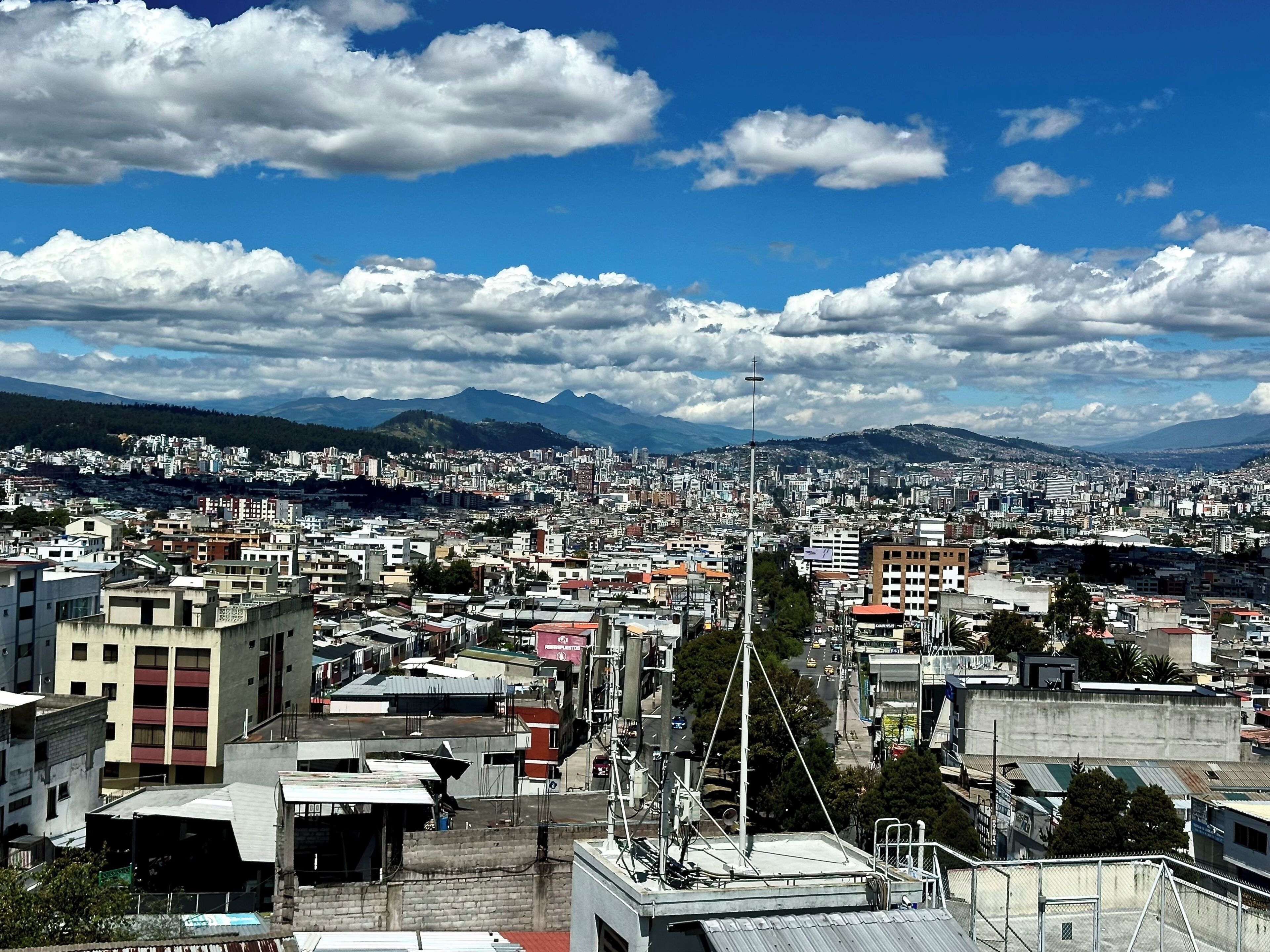 Une vue urbaine mêlant des immeubles résidentiels et commerciaux, un lampadaire bien visible et des montagnes lointaines sous un ciel partiellement nuageux.