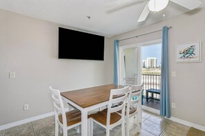 Een eethoek met een houten tafel en witte stoelen, een flatscreen tv aan de muur en uitzicht op de omgeving via een glazen deur.
