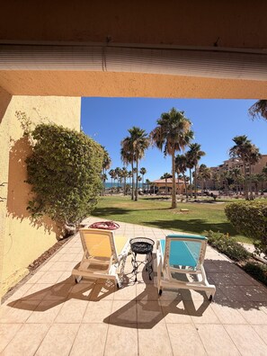 A patio with two lounge chairs and a small table, overlooking a lawn with palm trees and a building in the background. - Princesa Resort D-105 A Ground Floor Ocean view Condo (Puerto Peñasco)