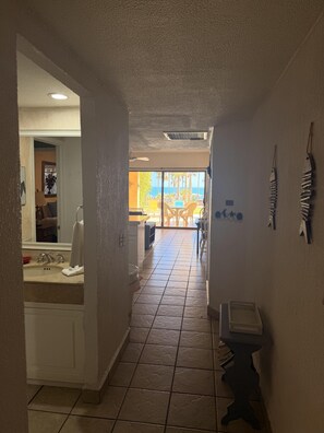 A hotel corridor with a bathroom on the left, a dining area with chairs and a table in the center, and a view of the ocean through large windows. - Princesa Resort D-105 A Ground Floor Ocean view Condo (Puerto Peñasco)