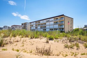 A multi-story hotel with balconies, an American flag, and a sandy foreground with sparse vegetation. - OceanfrontStudio-Sleeps2-Pool-FreeParking (Ocean City)