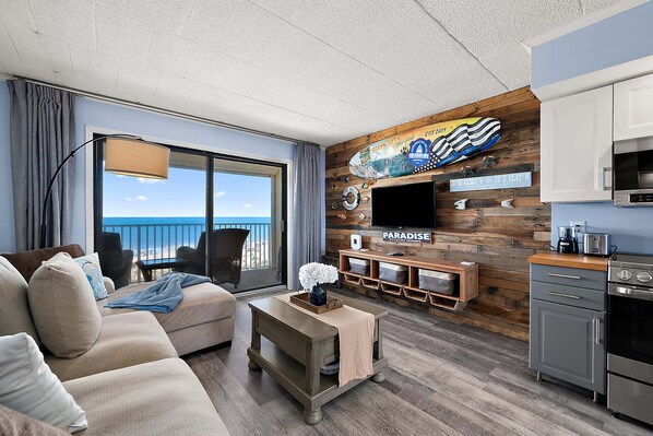 A modern living room with a large window, a flat-screen TV mounted on a wooden wall, a sofa, a coffee table, and a kitchenette. - OceanfrontStudio-Sleeps2-Pool-FreeParking (Ocean City)