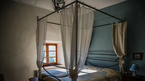 A four-poster canopy bed with a floral bedspread, a bedside table with a lamp, and a window with curtains.