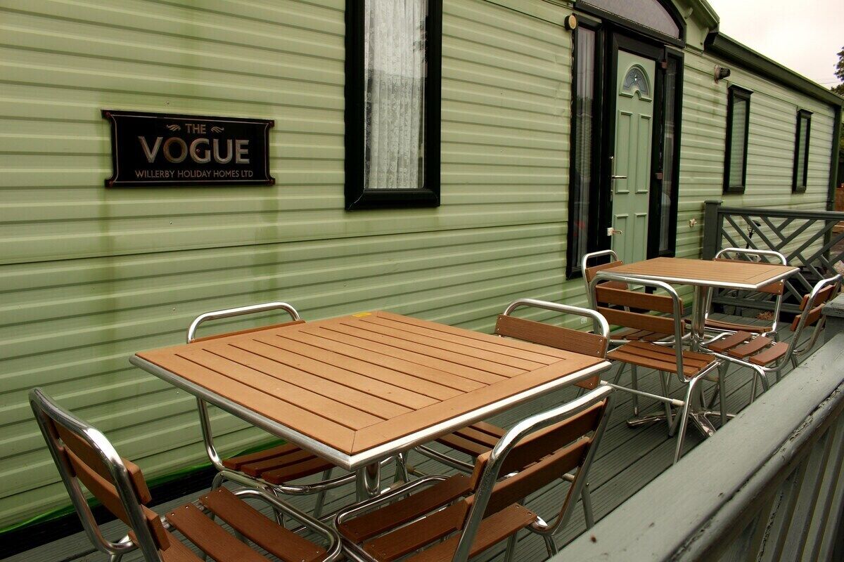 Pared exterior verde con un letrero que dice 'The Vogue Willerby Holiday Homes Ltd.' y varias mesas y sillas de madera dispuestas en una terraza.