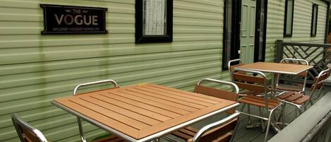Pared exterior verde con un letrero que dice 'The Vogue Willerby Holiday Homes Ltd.' y varias mesas y sillas de madera dispuestas en una terraza.