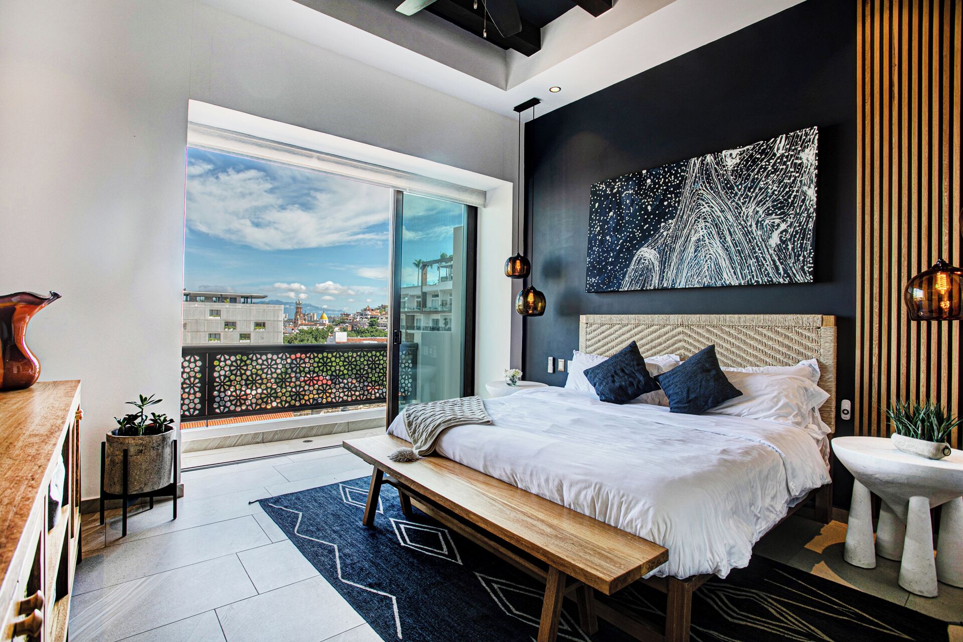 A modern bedroom with a large bed, a bench, a view of the city, and a cityscape painting.