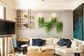 Ein modernes Wohnzimmer mit einer Couch, einem Couchtisch und einem Fernseher. Es gibt Regale mit Pflanzen und ein großes Gemälde an der Wand.