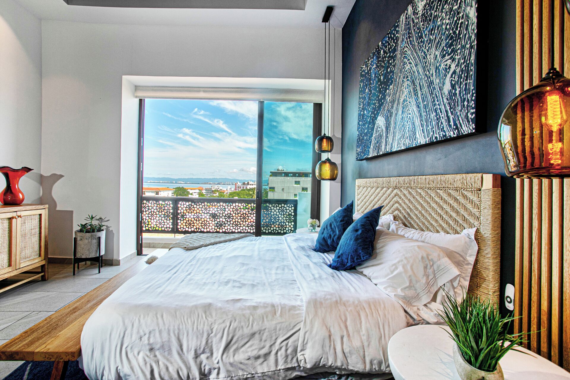 A modern bedroom with a large bed, a view of the city through a balcony, and a cityscape painting on the wall.