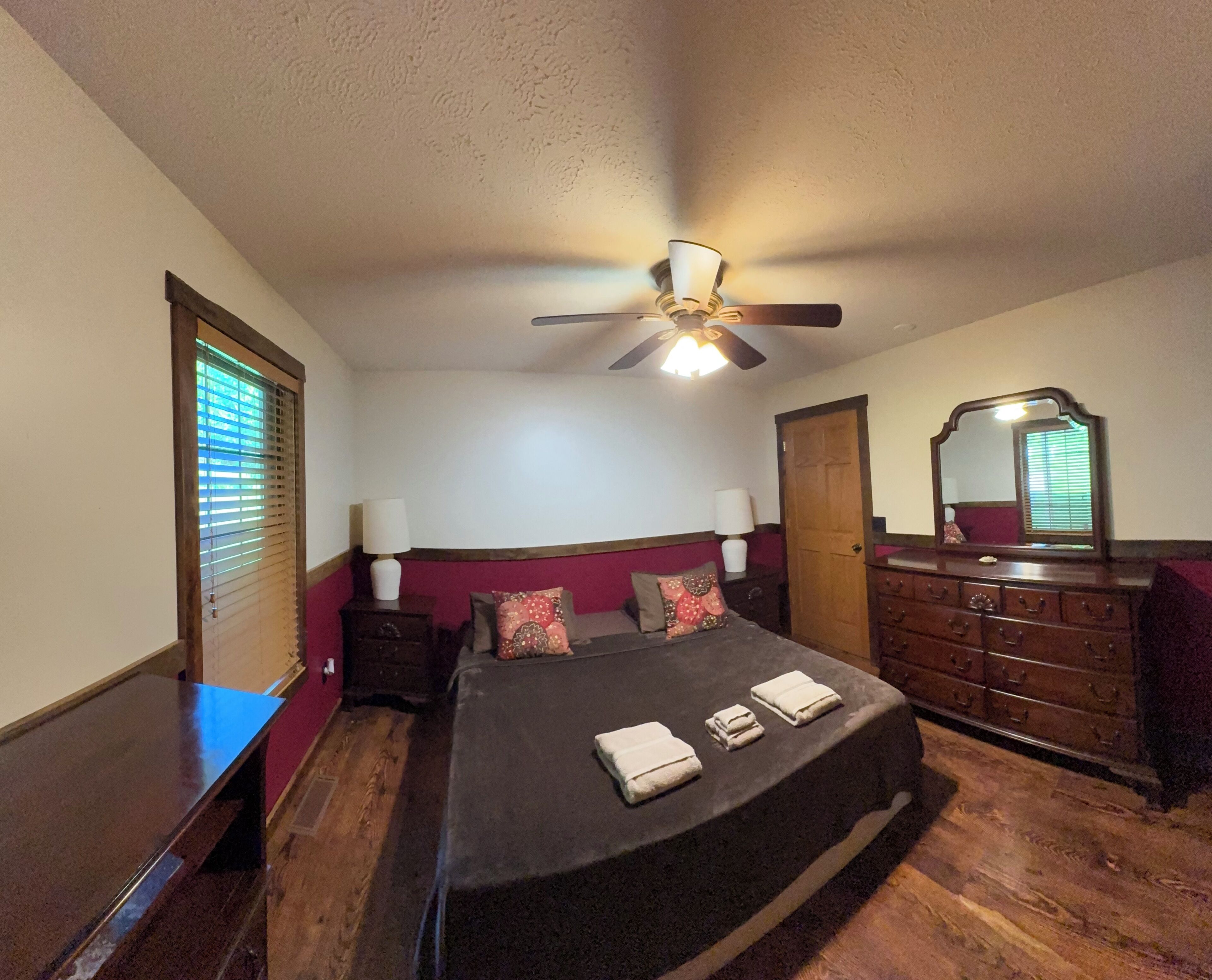 Une chambre à coucher comprenant un lit, une commode, un miroir, un ventilateur de plafond et une fenêtre avec des stores.