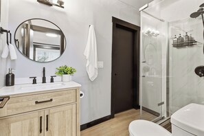 Een moderne badkamer met een houten wastafelmeubel, een ronde spiegel, een witte wasbak, een douchecabine van glas en een donkere deur.