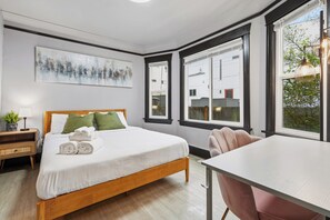 Een slaapkamer met een bed, nachtkastje, lamp en uitzicht op gebouwen door grote ramen.