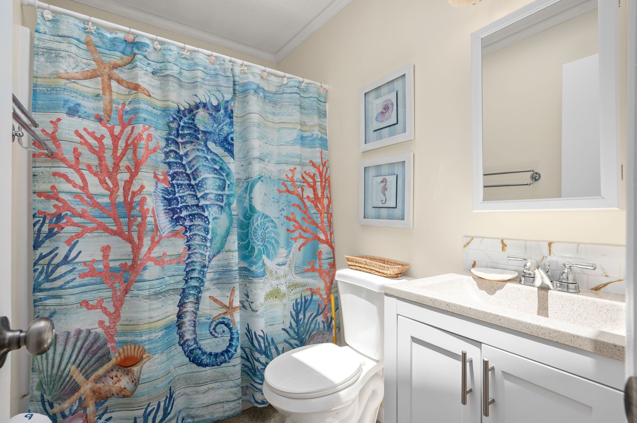 A bathroom with a seashore-themed shower curtain, a white toilet, a sink with a mirror, and framed pictures on the wall.