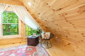 Eine Holzhütte mit einem großen Fenster, einem roten Teppich mit Muster, einer Holzkiste und einem Korbstuhl.