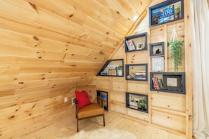 A cozy attic room with wooden walls, a leather chair, and a bookshelf filled with books and framed photos. - New Luxury Smoky Mountain Cabin, Hot Tub, Fire Pit, Mountain Views & Easy Access (SEVIERVILLE)