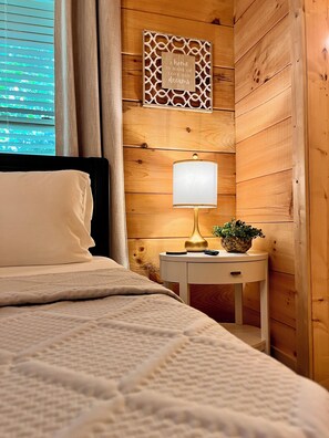 Ein Schlafzimmer mit einem Bett, einem Nachttisch mit Lampe, einem Wanddekor und einem Fenster mit Vorhängen.