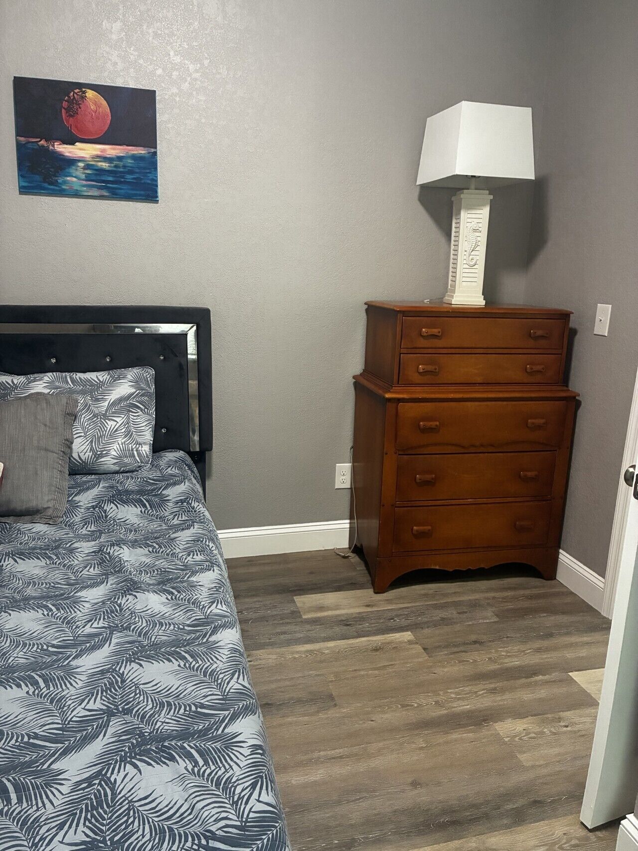 A bedroom with a bed, a dresser, a nightstand, a lamp, and a painting on the wall.