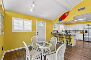 A yellow-walled room with a glass-top dining table, white wicker chairs, a white door, and a kitchen area with white cabinets and appliances. - 2BR, 2BA Beach House w/ Private Pool 11-Minute Walk to Shoreline (Panama City Beach)